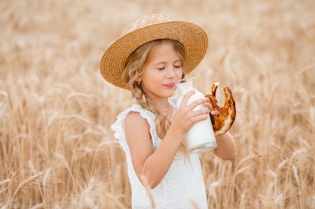 La piccola ragazza bionda nel campo di grano mangia il pane e beve il latte