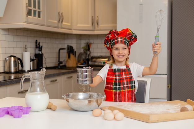 La piccola ragazza bionda sorridente felice in un costume da chef impasta la pasta per la cottura di Natale in cucina