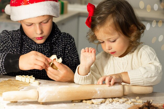 La piccola ragazza carina 2-4 con un fiocco rosso e il ragazzo 7-10 con un berretto natalizio preparano biscotti di pan di zenzero nella cucina di Capodanno.