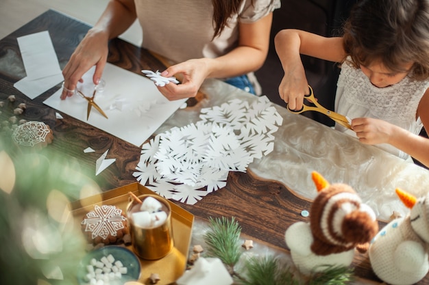 La piccola ragazza carina e la giovane bella donna hanno tagliato i fiocchi di neve da carta bianca. Pan di zenzero e cacao con marshmallow. Il concetto di preparazione per il nuovo anno e il Natale.