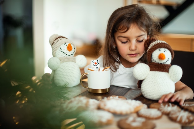 La piccola ragazza carina gioca con i pupazzi di neve lavorati a maglia e mangia il pan di zenzero e beve cacao con marshmallow.