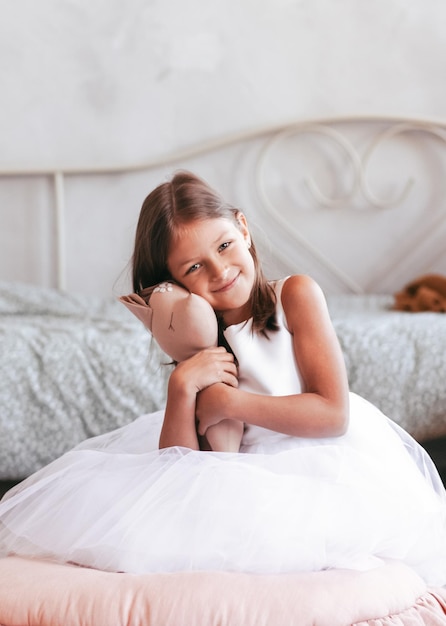 La piccola ragazza carina sorridente in un bellissimo vestito bianco è seduta con un peluche nelle sue mani