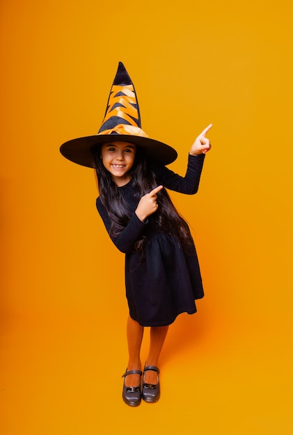 La piccola ragazza caucasica allegra in un costume da strega di Halloween punta il dito di lato.