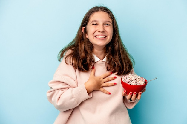 La piccola ragazza caucasica che tiene una ciotola di cereali isolata su sfondo blu ride ad alta voce tenendo la mano sul petto.