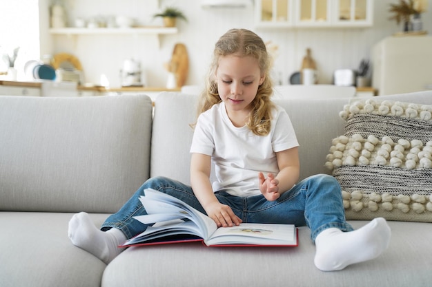 La piccola ragazza caucasica è seduta sul divano e sta leggendo il libro delle fiabe. Il bambino sta studiando a casa in quarantena. Il bambino in età prescolare sta imparando a leggere. Tempo libero e studio in una casa accogliente in quarantena.