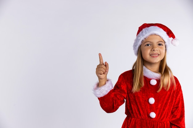 La piccola ragazza caucasica in cappello della Santa e costume rosso con un bel sorriso punta il dito. Isolato