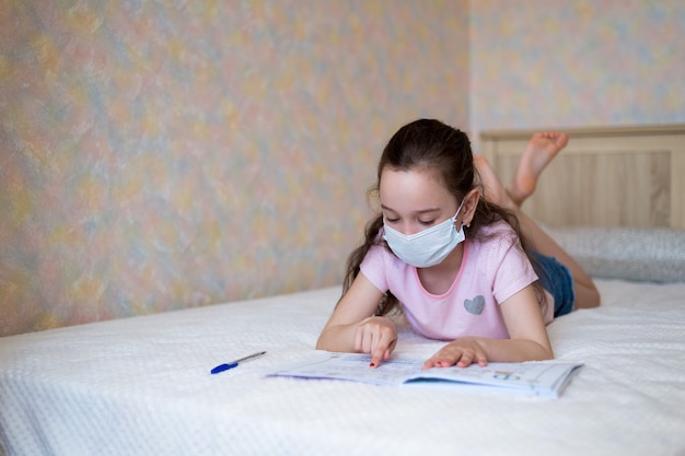 La piccola ragazza caucasica in una maschera protettiva scrive in un taccuino durante la quarantena