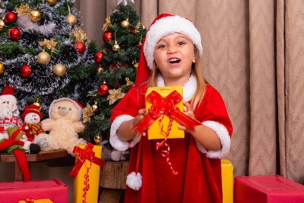 La piccola ragazza caucasica sveglia si è vestita per natale a casa che tiene un regalo.