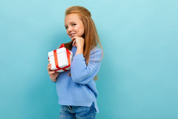 La piccola ragazza caucasica tiene una scatola bianca con il regalo e ha molte emozioni isolate sul blu