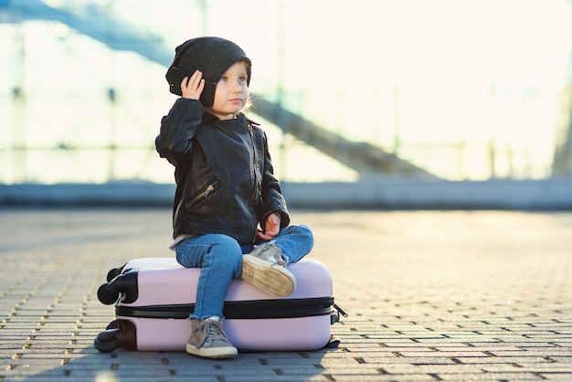 La piccola ragazza del viaggiatore si siede sulla valigia rosa e parla sullo smartphone vicino all'aeroporto contro il tramonto.