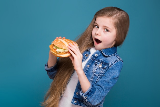 La piccola ragazza graziosa in giacca di jeans con lunghi capelli castani tiene un hamburger