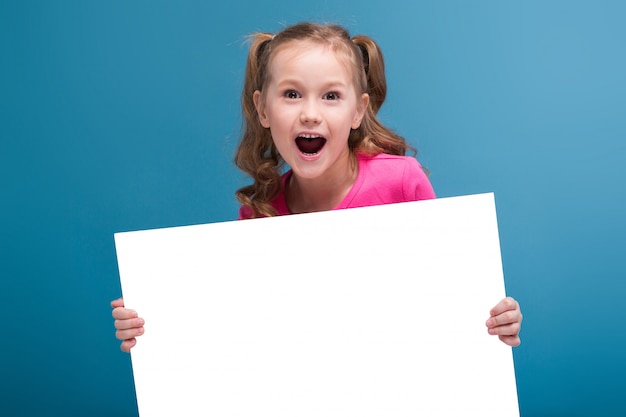 La piccola ragazza sveglia attraente in camicia rosa con la scimmia ed i pantaloni blu tengono il manifesto vuoto