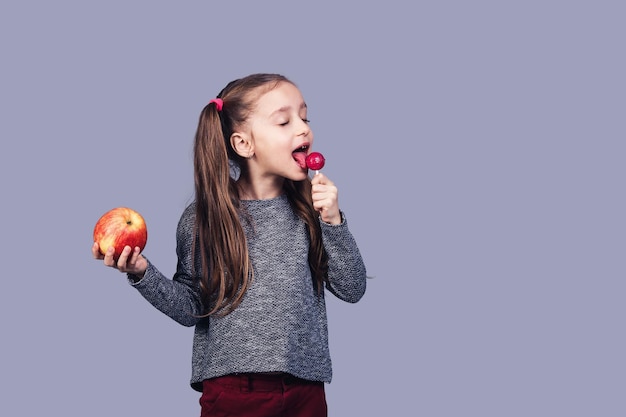 La piccola ragazza sveglia lecca un lecca-lecca e tiene una mela