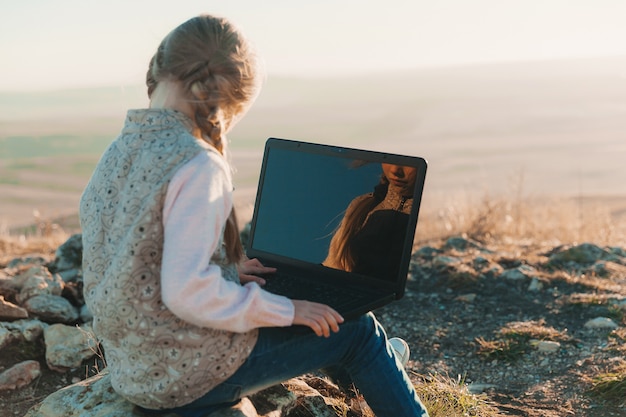 La piccola ragazza teenager felice si siede su una collina con un computer portatile. Nomadi digitali