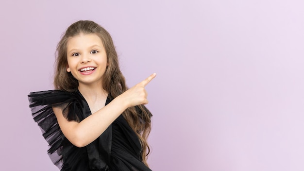 La piccola signora indica il tuo annuncio Una bella bambina con i capelli ricci in un vestito nero su sfondo rosa isolato Copia spazio