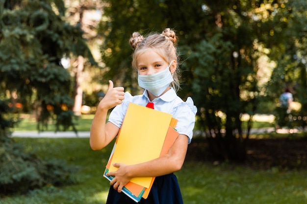 La piccola studentessa con una maschera medica si siede nel parco sull'erba e si diverte.