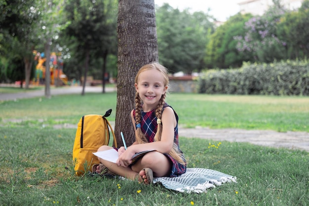 La piccola studentessa sorridente fa i compiti seduta sotto l'albero verde nel parco cittadino