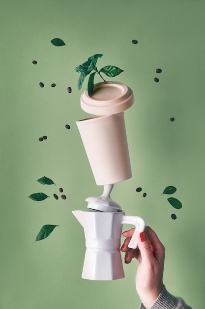 La piramide zero del caffè residuo di equilibratura ha tenuto dalla mano femminile sul fondo verde della menta. Caffettiera in ceramica per caffè espresso e tazza da caffè in bambù riutilizzabile eco-compatibile. Chicchi di caffè e foglie di piante.