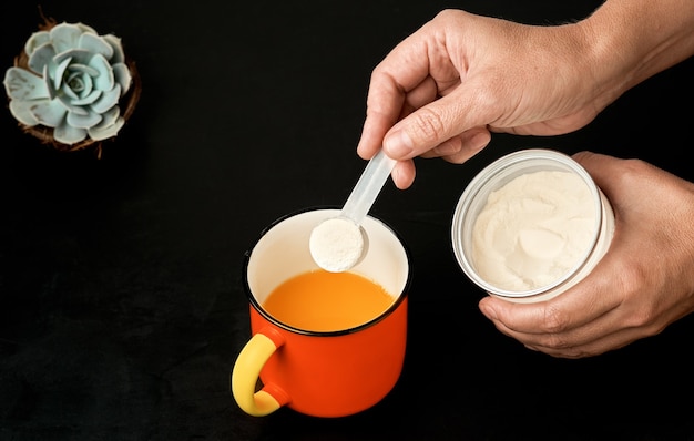 La polvere di proteine del collagene da un misurino di plastica mani femminili versare in una tazza con succo sul nero