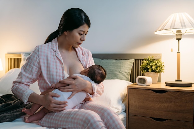 La premurosa mamma asiatica per la prima volta seduta al capezzale guarda la sua neonata nelle sue coccole mentre allatta il latte materno in camera da letto di notte.