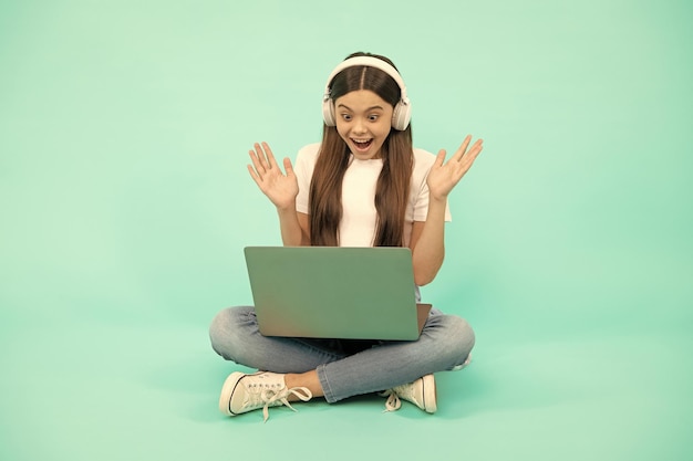 La ragazza adolescente sorpresa usa il laptop wireless per la videochiamata o ascolta il webinar in cuffia acquista online