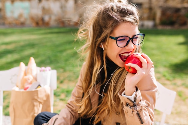 La ragazza affamata divertente con appetito morde la mela rossa che si siede nel parco dopo un viaggio di shopping. Attraente giovane donna che mangia voltarsi sullo sfondo della natura sfocata. Pranzo al parco, comprando cibo