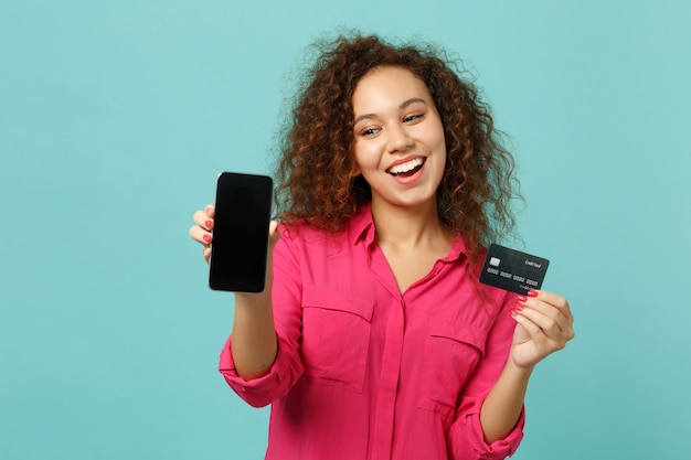 La ragazza africana allegra in abbigliamento casual tiene il telefono cellulare con la carta bancaria di credito dello schermo vuoto in bianco isolata su fondo blu del turchese. Concetto di stile di vita di emozioni sincere della gente. Mock up copia spazio.