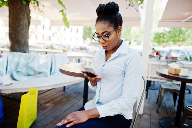 La ragazza afroamericana indossa gli occhiali con il telefono cellulare seduto al caffè all'aperto