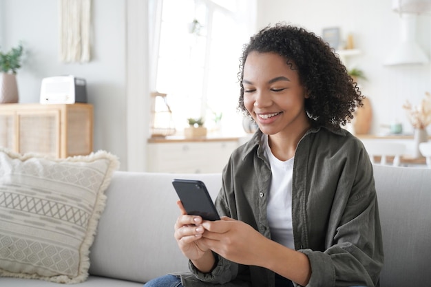 La ragazza afroamericana riccia si diverte con il telefono a casa e sorridente Concetto di comunicazione