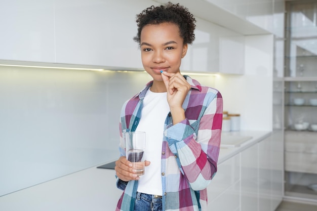 La ragazza afroamericana tiene il bicchiere d'acqua della pillola che assume l'integratore alimentare di vitamine per il benessere