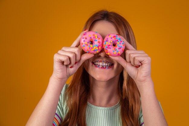 La ragazza alla moda con i capelli lunghi posa positivamente, tenendo ciambelle rosa fresche con polvere pronte per godersi i dolci.