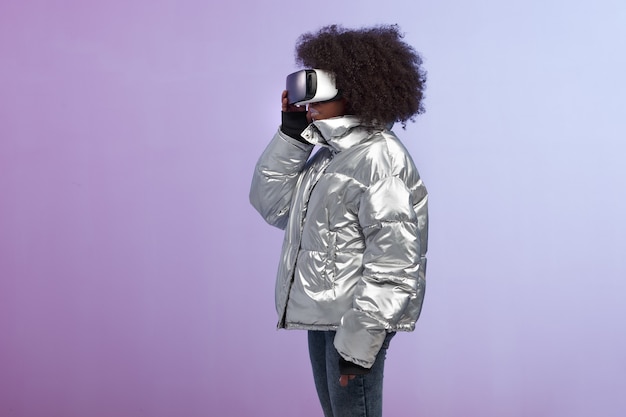 La ragazza alla moda dai capelli castani riccia vestita con una giacca color argento utilizza gli occhiali per realtà virtuale in studio su sfondo al neon.