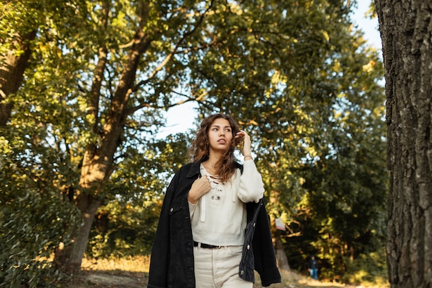 La ragazza alla moda dai capelli ricci in abiti casual eleganti in denim autunnale cammina sulla natura in campagna