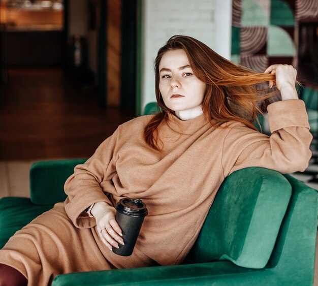 La ragazza alla moda si siede in un caffè al tavolo e beve il caffè. Caffè per andare in una tazza di cartone. Donna con i capelli rossi in un abito caldo beige in un'atmosfera accogliente. Interni moderni. Passatempo calmo e piacevole.