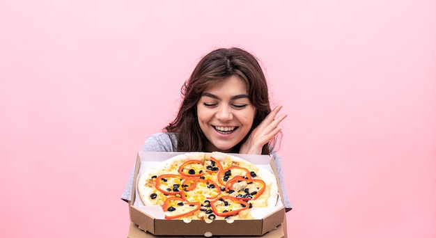 La ragazza allegra guarda in modo appetitoso la pizza appena sfornata in una scatola per la consegna su uno sfondo rosa