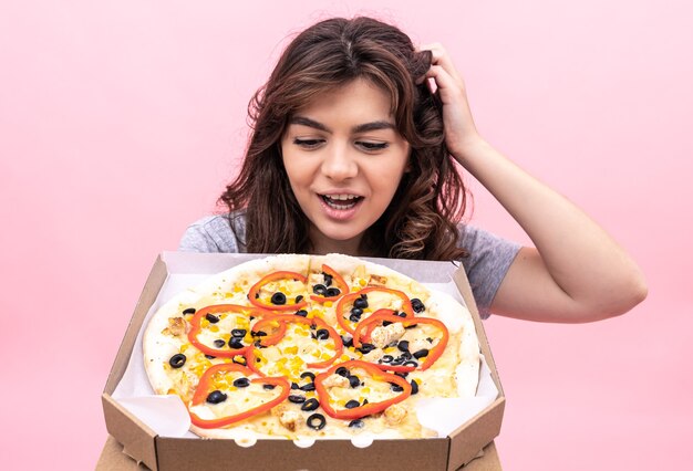 La ragazza allegra guarda in modo appetitoso la pizza appena sfornata in una scatola per la consegna su uno sfondo rosa.