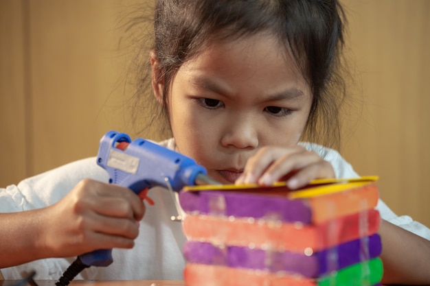 La ragazza asiatica del bambino che incolla il gelato colorato attacca dalla pistola per colla elettrica della colata a caldo. I bambini si divertono a fare casa su un progetto artigianale.