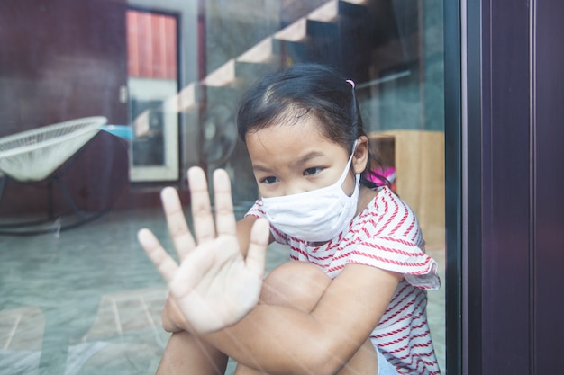 La ragazza asiatica del bambino che indossa la maschera di protezione guarda fuori dalla finestra e resta a casa in quarantena dal coronavirus Covid-19 e dall'inquinamento atmosferico pm2.5.