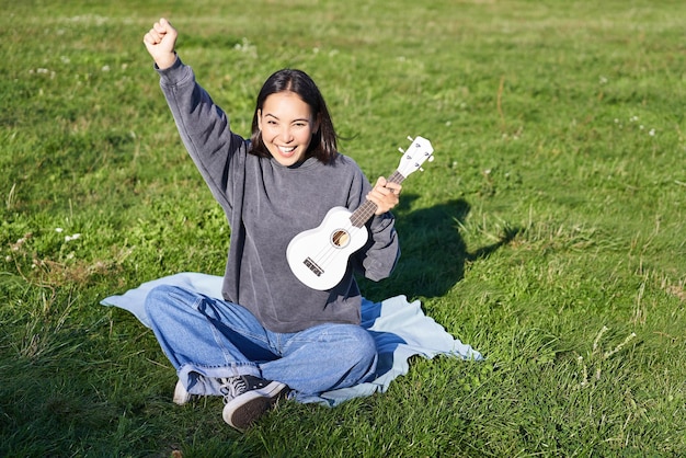 La ragazza asiatica eccitata si siede nel parco con l'ukulele suona lo strumento e si sente felice ottimista momenti positivi e concetto di persone