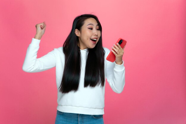 La ragazza asiatica felice eccitata tiene il telefono sentendosi euforica dal gioco online mobile che celebra la vittoria
