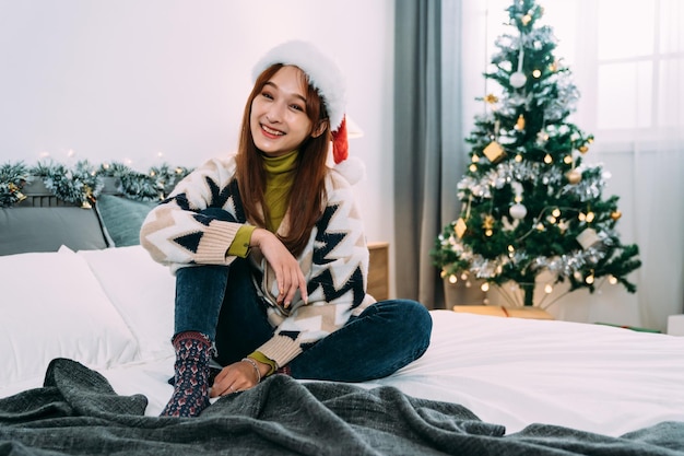 la ragazza asiatica sveglia che si siede sul letto con un cappello festivo sta sorridendo alla macchina fotografica in una camera da letto luminosa decorata di Natale a casa.