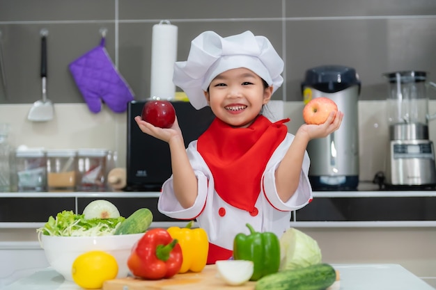 La ragazza asiatica sveglia indossa l'uniforme da chef con un sacco di verdure sul tavolo nella stanza della cucina Prepara cibo da mangiare a cena Tempo divertente per i bambini