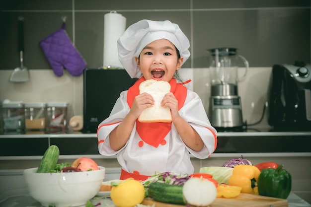 La ragazza asiatica sveglia indossa l'uniforme da chef con un sacco di verdure sul tavolo nella stanza della cucina Prepara cibo da mangiare a cena Tempo divertente per i bambini