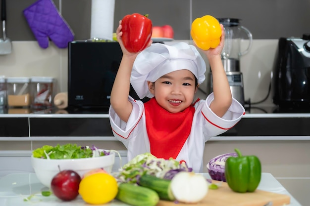 La ragazza asiatica sveglia indossa l'uniforme da chef con un sacco di verdure sul tavolo nella stanza della cucina Prepara cibo da mangiare a cena Tempo divertente per i bambini