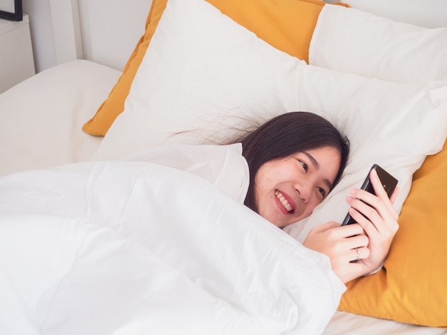 La ragazza asiatica sveglia sul letto e giocando con il telefono cellulare