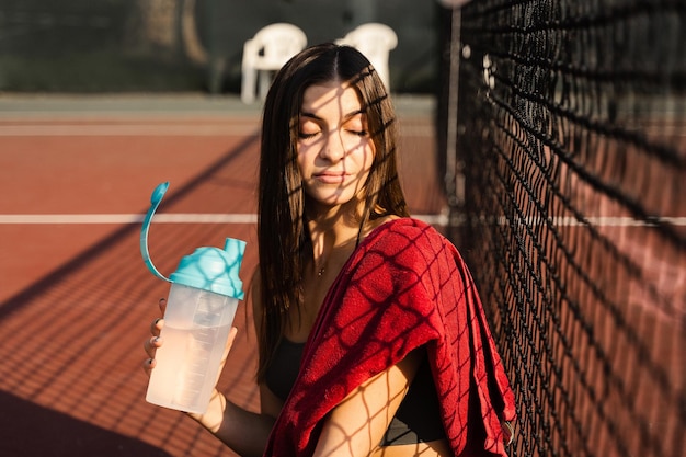 La ragazza atletica beve l'acqua da una bottiglia Riposa dopo l'allenamento sul campo da tennis