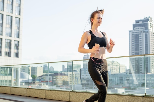 La ragazza atletica corre per allenarsi al mattino sullo sfondo della città e ascolta musica