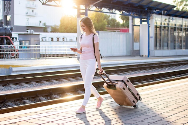 La ragazza attraente è in piedi con i bagagli alla stazione e aspetta il treno