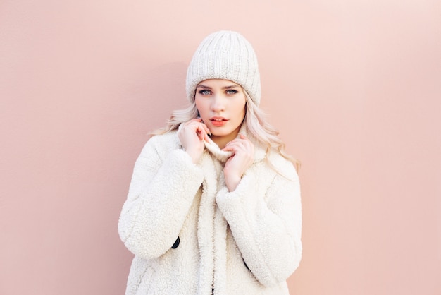 La ragazza bionda felice in inverno bianco copre la posa contro una parete rosa.