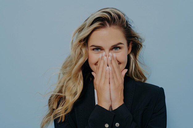 La ragazza bionda in cappotto nero copre la bocca con le mani che esprimono sorpresa e gioia in piedi su sfondo blu